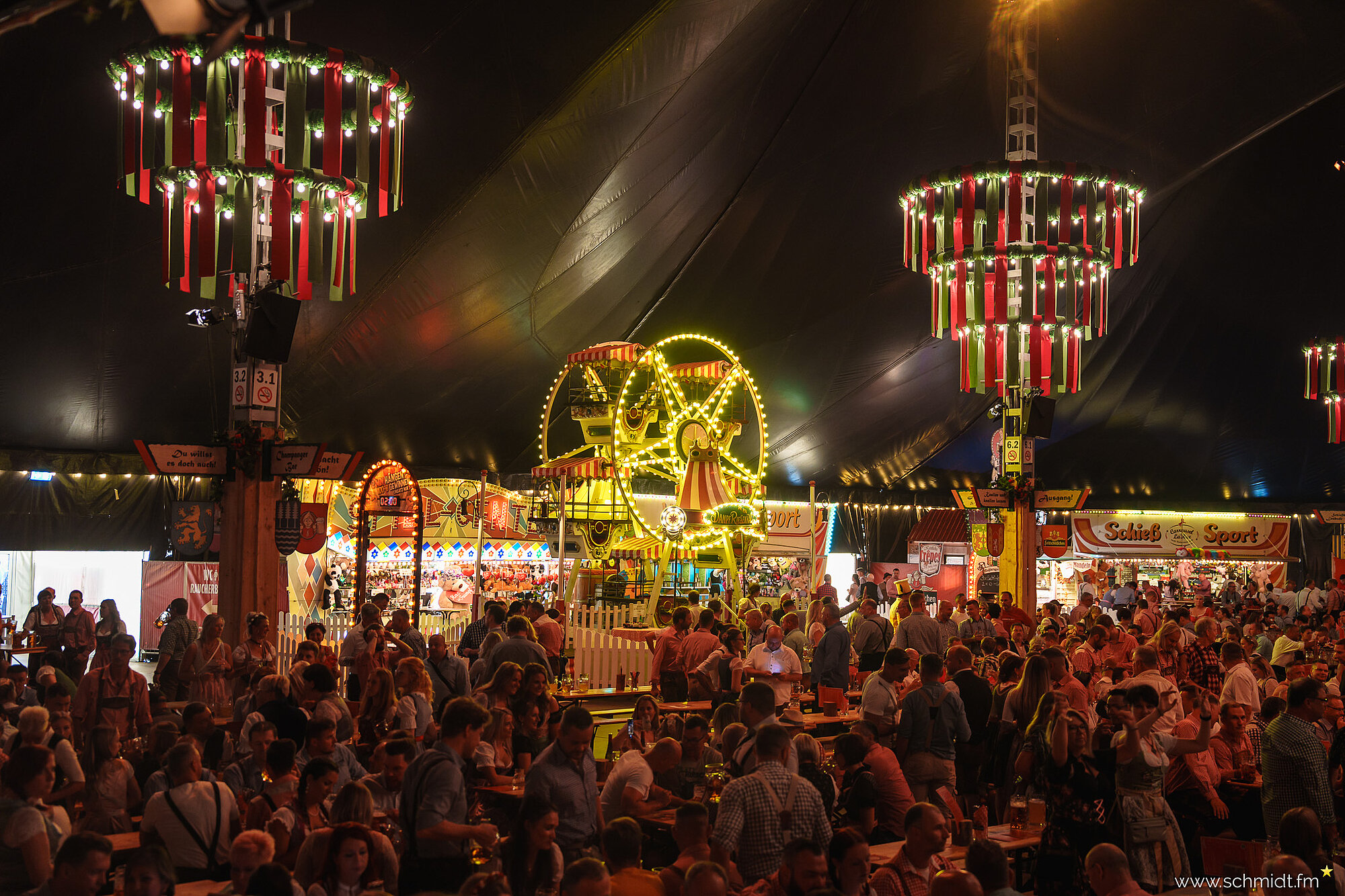 estlich dekoriertes Bierzelt mit Menschen in Dirndl und Lederhosen, die an langen Holztischen sitzen. Im Hintergrund das leuchtende historische Riesenrad